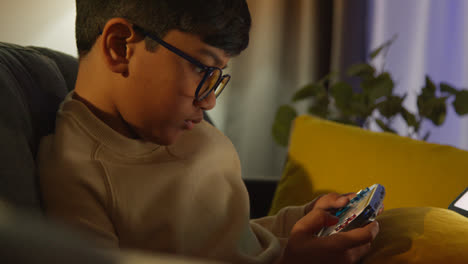 Young-Boy-Sitting-On-Sofa-At-Home-Playing-Game-Or-Streaming-Onto-Handheld-Gaming-Device-At-Night-4